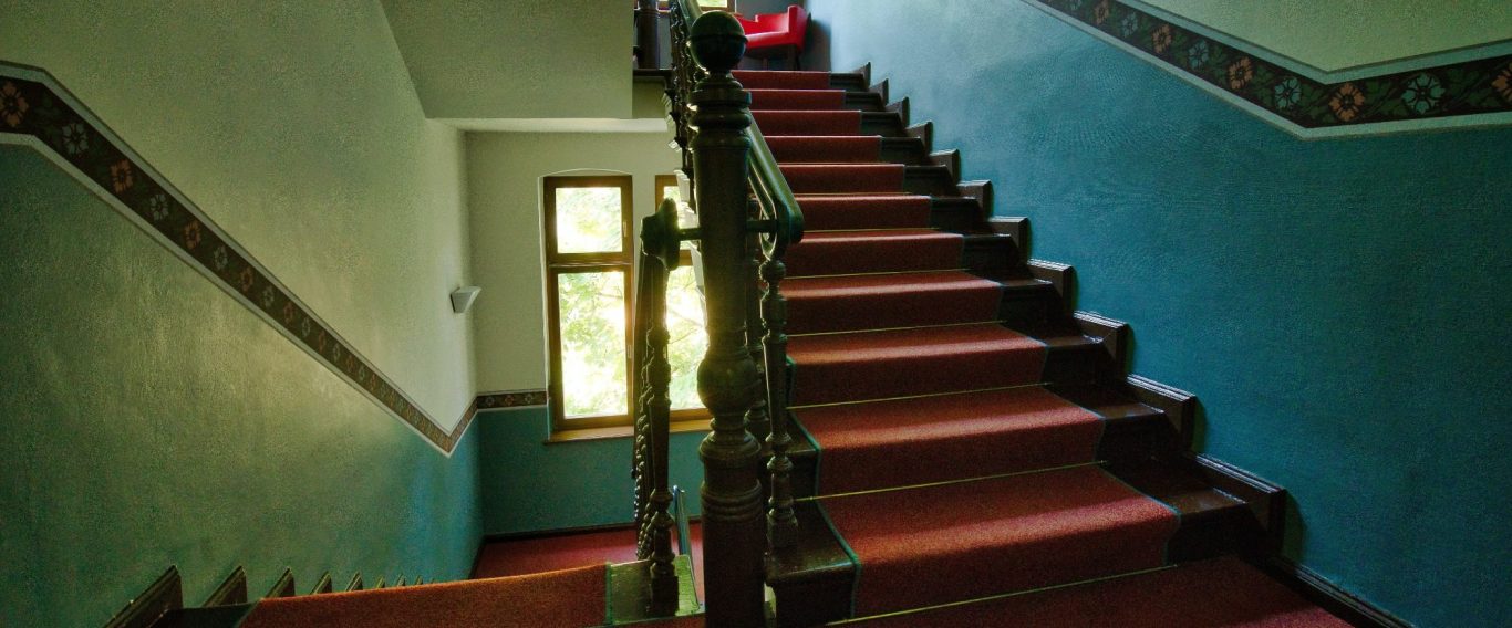 Treppenhaus mit grünblauen Wänden, Holzgeländer und einem roten Objekt auf der Treppe.