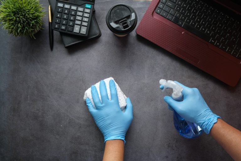 Hände in blauen Handschuhen reinigen einen Tisch mit Putzmittel und Tuch.